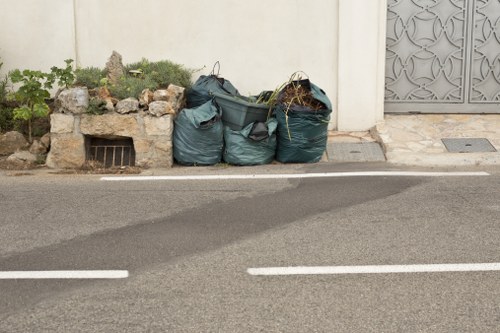 In the foreground, three dark green rubbish recycling bags are placed on the pavement, slightly leaning against a low stone wall with some small plants and rocks visible on top. The bags appear to be made of plastic, with a textured surface, and are filled with waste materials, causing them to bulge and form irregular shapes. Behind the bags, there is a plain cream-colored wall that extends across the background, with a small rectangular vent near the base of the wall. To the right, apart from the bags, is a section of decorative metal fencing or gate with a geometric pattern, suggesting an entrance or boundary for a property. The scene is lit by natural daylight, creating soft shadows on the pavement, which has a dark asphalt surface marked with white lines for vehicle parking. The location appears to be a residential or commercial street, possibly within West Ham, an area associated with the postcode E13, with the waste disposal activity characteristic of rubbish removal services offered by Commercial Waste West Ham.