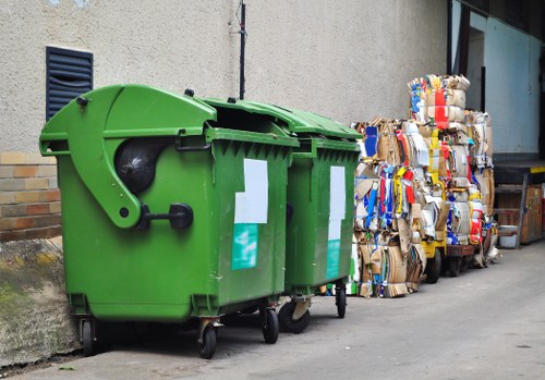 A pair of large, green wheelie bins made from durable plastic with smooth, shiny surfaces are positioned on a concrete pathway adjacent to a rough-textured, beige building wall. Each bin features a black lid with a curved edge and a wheel for mobility, with the lids slightly open revealing the dark interior. The bins are situated next to a stack of tightly packed cardboard and paper waste, which is bound with tape and plastic strapping. The cardboard waste includes flattened boxes of various sizes, with some displaying printed graphics and logos, predominantly in shades of brown, white, and coloured packaging remnants. The setting appears to be outdoors in a narrow alley or service area, with the bins and waste positioned close to the wall, suggesting cleaning or disposal activities typical for commercial waste collection in West Ham. The environment is lit with natural daylight, casting minimal shadows, and the background features a portion of a glass window reflecting the surroundings, with a small vent visible on the wall to the left. The scene effectively illustrates waste management practices related to rubbish collection for local businesses managed by Commercial Waste West Ham.