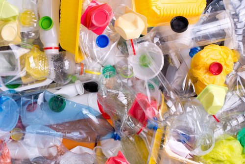 A close-up view of a large pile of discarded plastic waste, predominantly consisting of transparent PET bottles and various plastic caps in multiple colours such as red, green, blue, and yellow. The bottles, some with labels and others plain, are crumpled and piled on top of each other, with caps firmly attached or loose. The background shows a mixture of additional plastic debris, with a yellow object partially visible among the waste, indicating an accumulation likely found at a waste collection or disposal site near West Ham, east London. The textures vary from smooth and glossy surfaces of the plastic bottles to the matte finish of the caps, emphasizing the variety of plastic types involved. The overall scene reflects the environmental impact of plastic consumption, suitable for a commercial rubbish removal context handled by companies like Commercial Waste West Ham, specialising in the collection and disposal of waste for local businesses or industrial sites in the area.