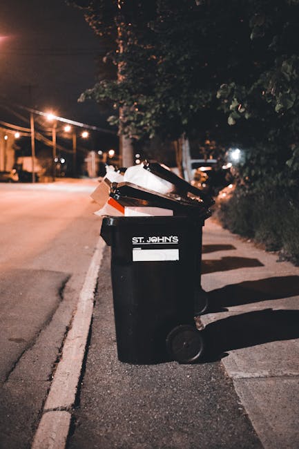 A black wheelie bin with the label 'St. John's' positioned on a pavement beside a street at night. The bin is open at the top, filled with various types of domestic rubbish, including cardboard boxes 