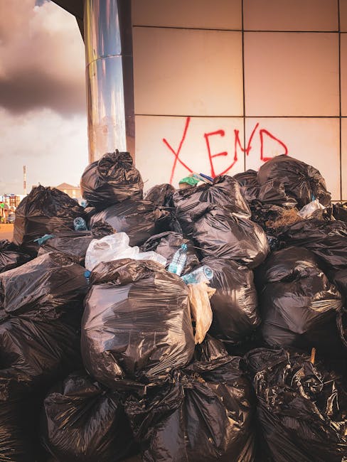 A large pile of black rubbish bags is stacked against a modern building with a reflective metal surface and beige tiled walls, where the word 'XEND' is spray-painted in red graffiti. Some bags are sli